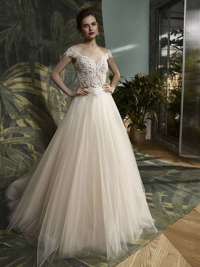 Code: Enzoani Blue - Kitara - IN STUDIO Romantic, dreamy, and sexy all in one -- this full-length A-line gown is modern bridal beauty at its best with illusion, off-the-shoulder cap sleeves on an unlined bodice of beaded, embroidered lace atop a flowing, soft tulle skirt. Striking crystal buttons trailing down the intricate illusion lace back provide the sweet and stunning finishing touch.Colour Options: Champagne/Nude  OR  Ivory/Nude  OR  Ivory/Ivory