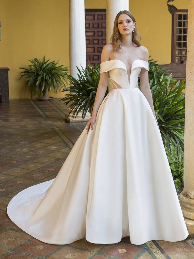 Code: Enzoani Blue Pearson This romantic-meets-modern bridal look is a true vision. This off-the-shoulder Mikado ball gown features a V-plunge illusion sweetheart neckline, contemporary cuts with illusion sheer side panels, and unique corset-style back with gorgeously designed boning and art deco vibes. It’s meant to contour the waist and emphasize the voluminous, chic skirt. Covered buttons cascade down from the corset back to the hem of the gown for that extra sweet touch.Colour Options: Ivory/Nude OR Ivory/Ivory