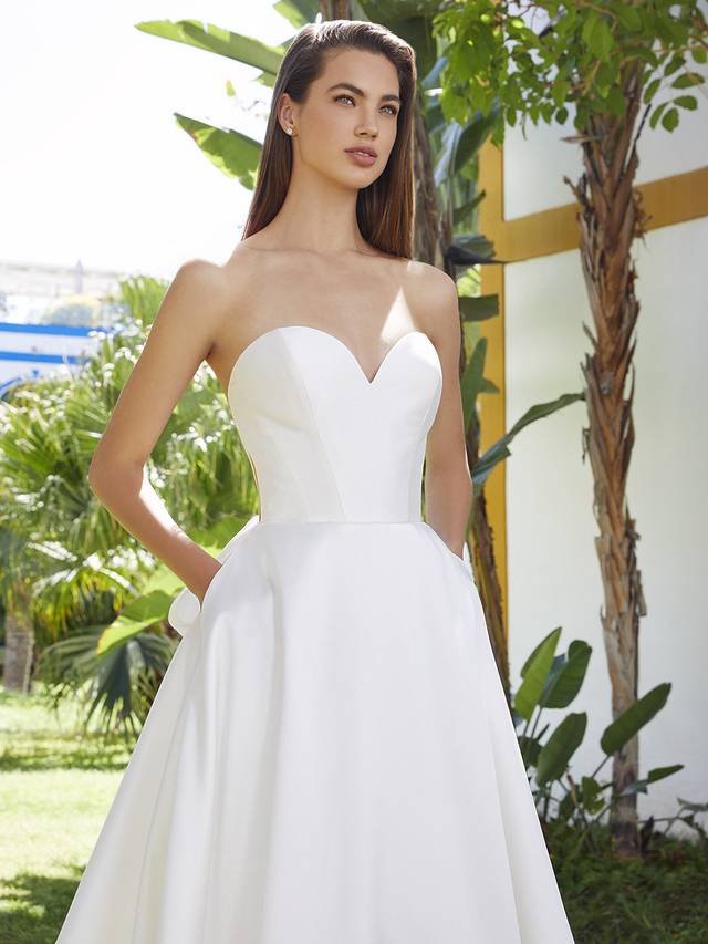 Code: Enzoani Love Bellarose Fall in love with BELLAROSE. The simple sweetheart neckline and statement back details are perfect. Along with a modern, illusion corset detail bodice, the dazzling drama continues with the showstopping bow detail on the back that complements delicate covered buttons trailing all the way down the back of the gown. And don’t forget – there are pockets! Colour Options: Ivory/Nude OR Ivory/Ivory