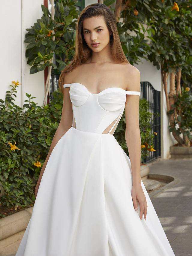 Code: Enzoani Love Beverly This lightweight matte Mikado gown is simple with a statement. Buttons cascade down the back of the entire dress to add delicate detailing and dimension to the regal, full circle A-line skirt. The illusion corset detail on the sides of the bodice along with a modern, updated sweetheart neckline create a flattering, princess-like silhouette. Sleek, thin off-the-shoulder sleeves and a peek-a-boo front slit provide even more subtly sexy, glamorous touches. Colour Options: Ivory/Nude OR Ivory/Ivory