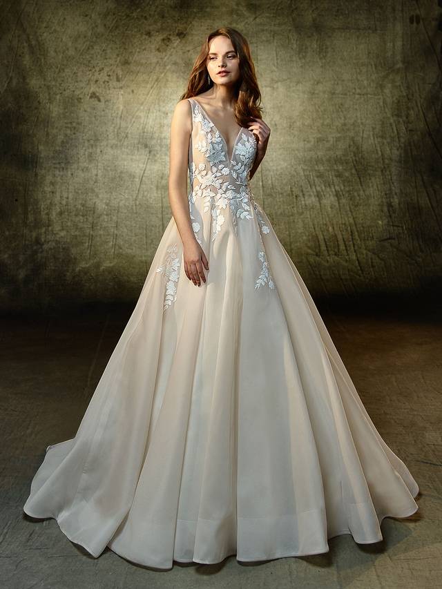 Code: Enzoani Blue Lennox Code: Enzoani Blue Lennox Simply sweet and divine, this full A-line gown is sure to captivate with embroidered floral lace that beautifully flows throughout the deep, unlined illusion V-neckline bodice and soft organza skirt. The unlined, low V-back complements the front as it continues with a sweet V-back embellished with delicate crystal buttons.Colour Options : Dark Ivory/Ivory OR Ivory/Ivory