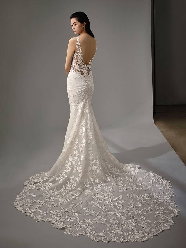 Code: Enzoani Blue - Malia Code: Enzoani Blue - Malia Become mesmerized with the Malia gown. Modern and sultry, this full length mermaid gown offers an enviable floral train. Delicate floral lace appliques are placed onto the bodice cascading down to the hips. Stretch georgette harmoniously compliments the lace as it is then ruched in the back of the gown. Finished with a playful low v-back, covered buttons, and an invisible zipper.Colour Options: Ivory