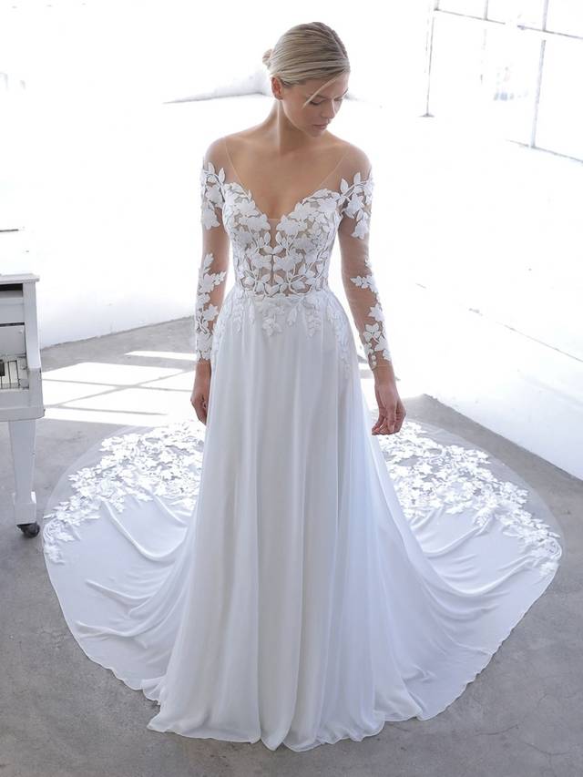 Code: Blue By Enzoani Nandana Code: Blue By Enzoani Nandana You will float down the aisle in this ethereal A-line gown! NANDANA features elegant, long tulle sleves with captivating 3D floral cotton lace and a lightweight chiffon skirt. The illusion tulle bodice gives you the appearance that this is an off-the-shoulder style. The back features covered buttons that lead to an invisible zipper finished with hand placed floral appliques along the train. Colour Options: Ivory/Nude OR Ivory/Ivory