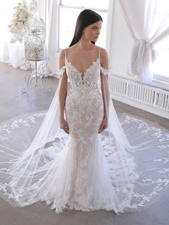 Code: Blue By Enzoani Olana (with cape) Code: Blue By Enzoani Olana (with cape) Like a gorgeous upgrade on our beloved Nadia gown. Combined with sparkling sequined Chantilly lace, the beloved thick embroidered floral lace stands out even more with figure-enhancing illusion side panels and the low, open illusion lace back. The detachable, off-shoulder lace and tulle split cape creates even more jaw-dropping drama. Colour Options: Ivory/Nude/Nude OR Ivory/Ivory/Nude OR Ivory/Sand/Nude OR Ivory/Ivory/Ivory
