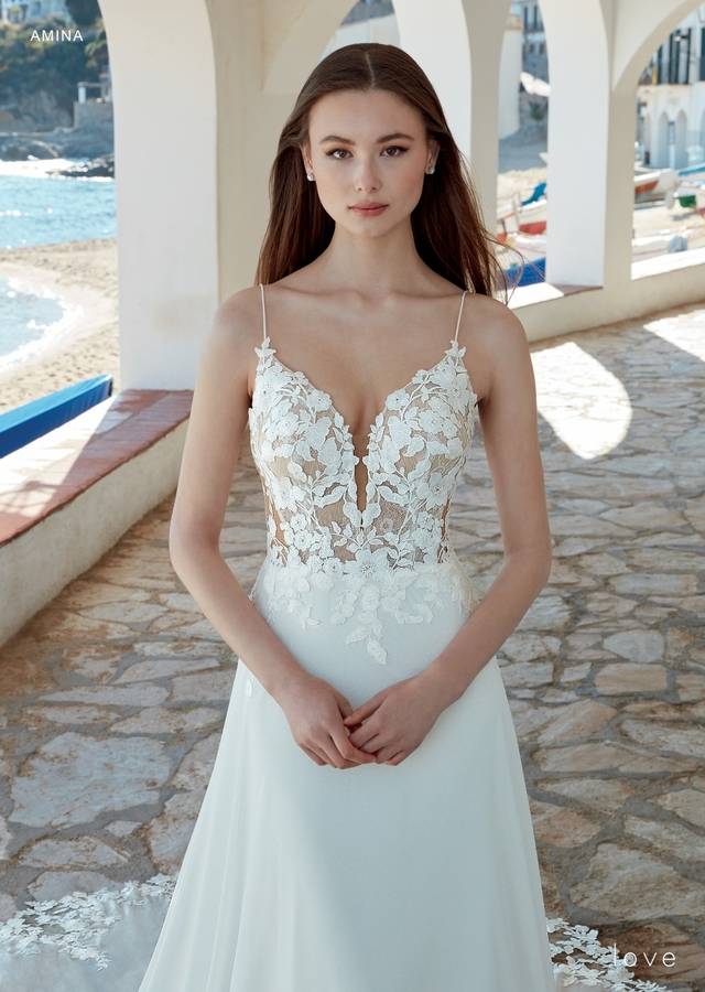 Code: Enzoani Love Amina - ARRIVING SOON Code: Enzoani Love Amina - ARRIVING SOON This gown has exquisite floral lace appliqués scattered delicately over the bodice, skirt and hem of the train for the most eye-catching effect. A plunging V-neck with spaghetti straps gives a sweet shape and support to the structured bodice, complementing a full circle skirt of floaty light chiffon with the extended hand-cut lace scalloped train. Colour Options: Ivory/Nude OR Ivory/Ivory