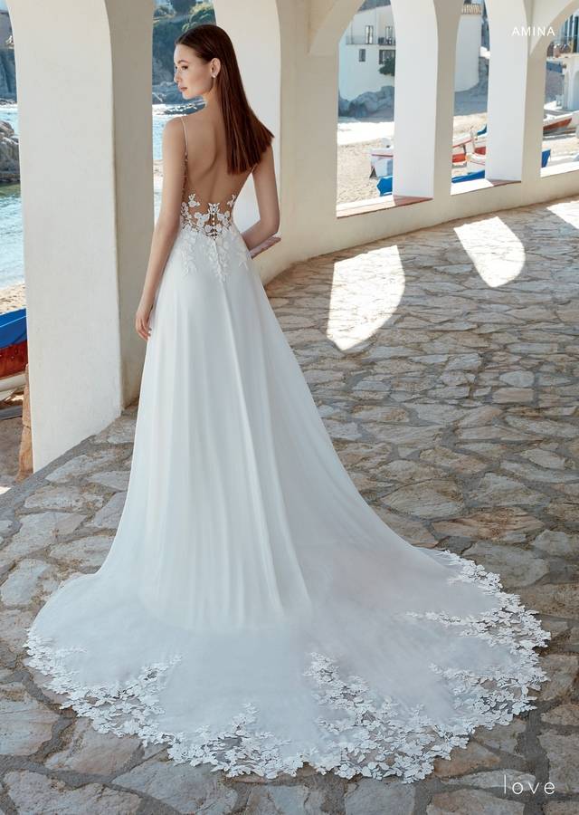 Code: Enzoani Love Amina - ARRIVING SOON Code: Enzoani Love Amina - ARRIVING SOON This gown has exquisite floral lace appliqués scattered delicately over the bodice, skirt and hem of the train for the most eye-catching effect. A plunging V-neck with spaghetti straps gives a sweet shape and support to the structured bodice, complementing a full circle skirt of floaty light chiffon with the extended hand-cut lace scalloped train. Colour Options: Ivory/Nude OR Ivory/Ivory