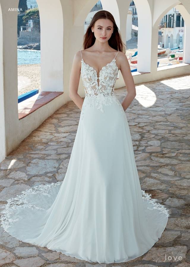 Code: Enzoani Love Amina - ARRIVING SOON Code: Enzoani Love Amina - ARRIVING SOON This gown has exquisite floral lace appliqués scattered delicately over the bodice, skirt and hem of the train for the most eye-catching effect. A plunging V-neck with spaghetti straps gives a sweet shape and support to the structured bodice, complementing a full circle skirt of floaty light chiffon with the extended hand-cut lace scalloped train.Colour Options: Ivory/Nude OR Ivory/Ivory