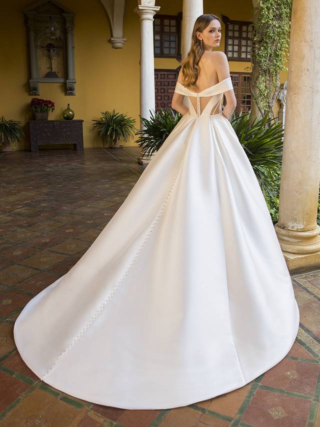 Code: Blue By Enzoani Pearson Code: Blue By Enzoani Pearson This romantic-meets-modern bridal look is a true vision. This off-the-shoulder Mikado ball gown features a V-plunge illusion sweetheart neckline, contemporary cuts with illusion sheer side panels, and unique corset-style back with gorgeously designed boning and art deco vibes. It’s meant to contour the waist and emphasize the voluminous, chic skirt. Covered buttons cascade down from the corset back to the hem of the gown for that extra sweet touch. Colour Options: Ivory/Nude OR Ivory/Ivory