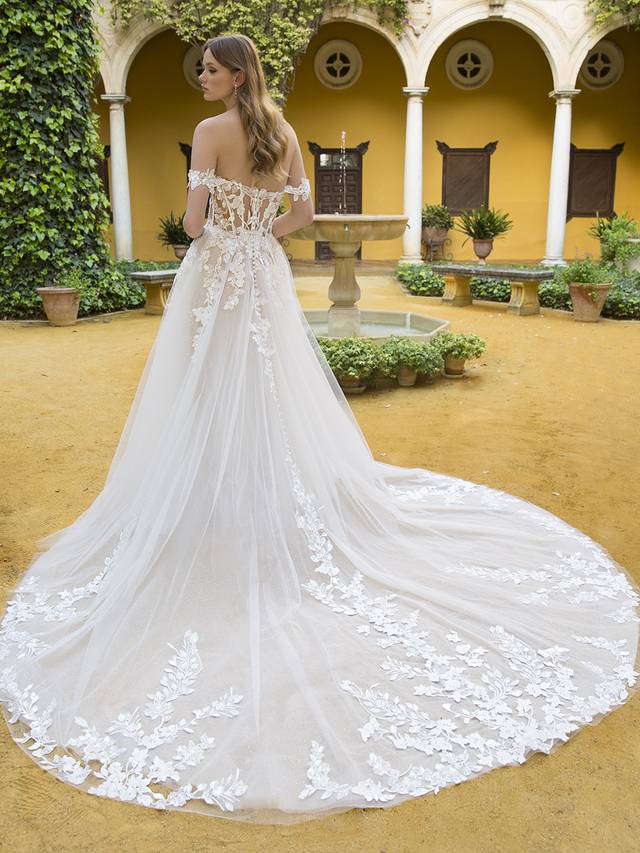 Code: Blue By Enzoani Petunia Code: Blue By Enzoani Petunia Sweet and ethereal, this lavish, full A-line bridal gown features a V-plunge illusion sweetheart neckline on a gorgeous shimmer beaded lace bodice and 3D floral embroidery off-the-shoulder straps. The whimsical appliques adorning the corset-style bodice gently trickle down to the lush skirt. This gown is perfect for the modern bride who wants both exquisite detailing and unique versatility. Ivory/Nude/Nude OR Ivory/Ivory/Ivory OR Ivory/Sand/Nude