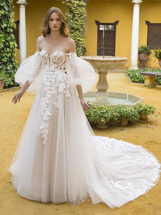 Code: Blue By Enzoani Petunia (With Sleeves) Code: Blue By Enzoani Petunia (With Sleeves) Sweet and ethereal, this lavish, full A-line bridal gown features a V-plunge illusion sweetheart neckline on a gorgeous shimmer beaded lace bodice, paired with romantic, detachable sheer tulle bishop sleeves and 3D floral embroidery off-the-shoulder straps. The whimsical appliques adorning the corset-style bodice gently trickle down to the lush skirt. This gown is perfect for the modern bride who wants both exquisite detailing and unique versatility. Ivory/Nude/Nude OR Ivory/Ivory/Ivory OR Ivory/Sand/Nude