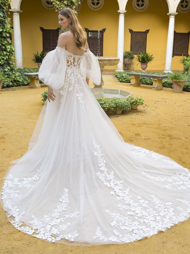 Code: Blue By Enzoani Petunia (With Sleeves) Code: Blue By Enzoani Petunia (With Sleeves) Sweet and ethereal, this lavish, full A-line bridal gown features a V-plunge illusion sweetheart neckline on a gorgeous shimmer beaded lace bodice, paired with romantic, detachable sheer tulle bishop sleeves and 3D floral embroidery off-the-shoulder straps. The whimsical appliques adorning the corset-style bodice gently trickle down to the lush skirt. This gown is perfect for the modern bride who wants both exquisite detailing and unique versatility. Ivory/Nude/Nude OR Ivory/Ivory/Ivory OR Ivory/Sand/Nude