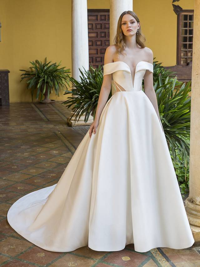 Code: Enzoani Blue Pearson Code: Enzoani Blue Pearson This romantic-meets-modern bridal look is a true vision. This off-the-shoulder Mikado ball gown features a V-plunge illusion sweetheart neckline, contemporary cuts with illusion sheer side panels, and unique corset-style back with gorgeously designed boning and art deco vibes. It’s meant to contour the waist and emphasize the voluminous, chic skirt. Covered buttons cascade down from the corset back to the hem of the gown for that extra sweet touch.Colour Options: Ivory/Nude OR Ivory/Ivory