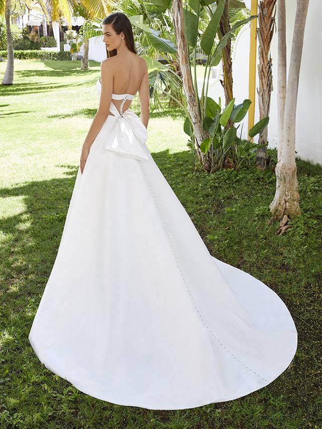Code: Love By Enzoani Bellarose Code: Love By Enzoani Bellarose Fall in love with BELLAROSE. The simple sweetheart neckline and statement back details are perfect. Along with a modern, illusion corset detail bodice, the dazzling drama continues with the showstopping bow detail on the back that complements delicate covered buttons trailing all the way down the back of the gown. And don’t forget – there are pockets! Colour Options: Ivory/Nude OR Ivory/Ivory